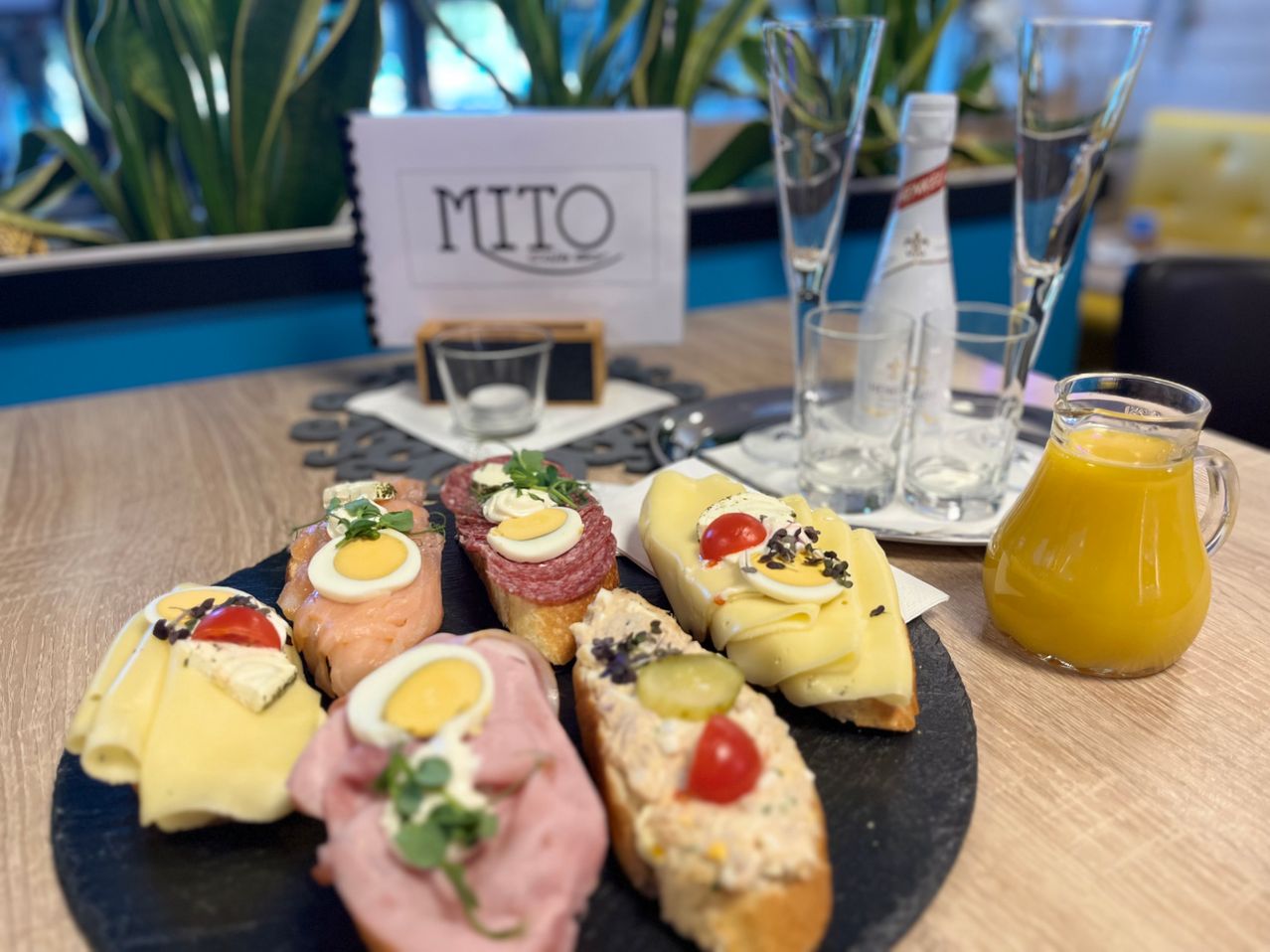 Auf einem Holztisch sind verschiedene Sandwiches zu sehen, darunter Schinken, Käse und Eierscheiben, begleitet von Gläsern, einer Flasche und einer Karaffe mit Orangensaft. Im Hintergrund ist ein Schild mit dem Wort MITO zu sehen.