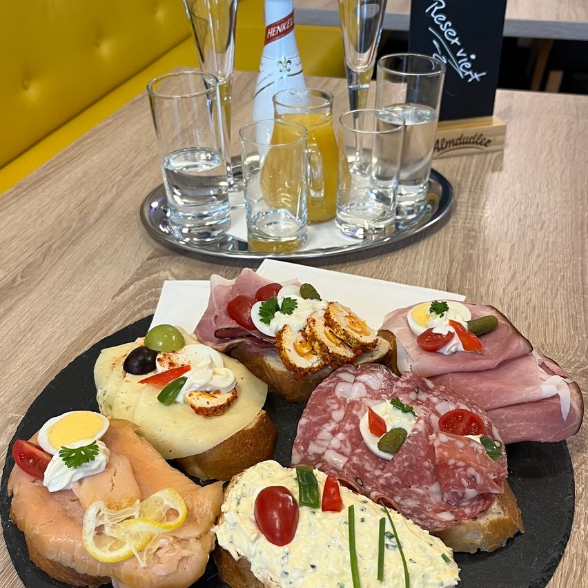 Ein Holztisch mit einem Tablett mit Gläsern und einer Karaffe mit Orangensaft und einem schwarzen Teller mit verschiedenen Sandwiches und Fleisch darauf.