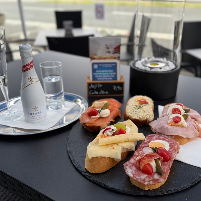 Ein Tisch mit einer Flasche Henkell Prosecco, einem Glas Wasser und einer Platte mit verschiedenen Sandwiches und Fleisch auf einer schwarzen Schieferplatte.