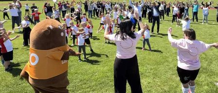 Eine große Gruppe von Menschen auf einem Feld mit einem Maskottchen, einige tragen weiße Hemden und heben ihre Hände. Bäume und Häuser sind im Hintergrund zu sehen.