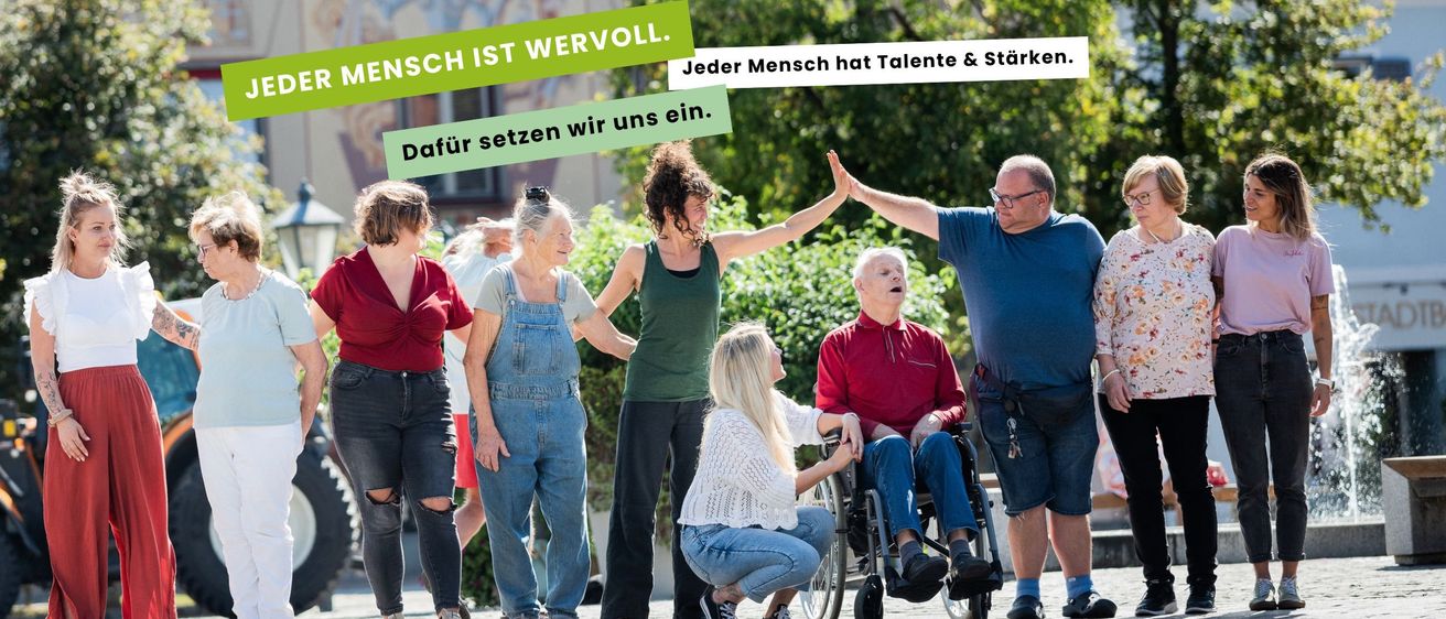 Eine Gruppe von unterschiedlichen Personen, darunter ältere Menschen und eine Frau im Rollstuhl, steht zusammen draußen. Sie lächeln und scheinen in ein Gespräch vertieft zu sein. Im Hintergrund befindet sich ein Gebäude mit einer Wandmalerei und Bäumen.