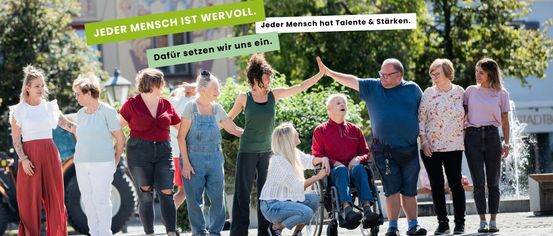 Eine Gruppe von unterschiedlichen Personen, darunter ältere Menschen und eine Frau im Rollstuhl, steht zusammen draußen. Sie lächeln und scheinen in ein Gespräch vertieft zu sein. Im Hintergrund befindet sich ein Gebäude mit einer Wandmalerei und Bäumen.