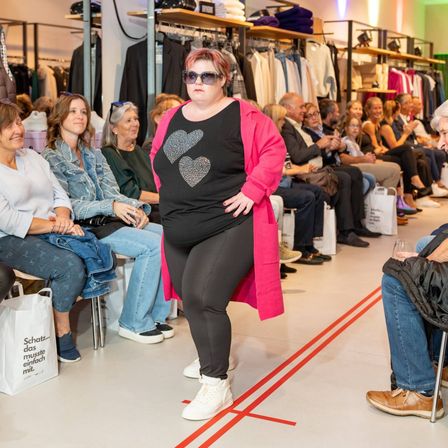 Eine Modenschau in einem Geschäft mit einem Publikum. Eine Frau in Schwarz und Pink geht auf einem markierten Pfad vor ihnen.