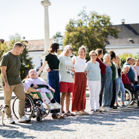 Eine Gruppe von Menschen, darunter einige in Rollstühlen, steht an einem sonnigen Tag vor einem Gebäude. Bäume und eine Statue sind im Hintergrund zu sehen.
