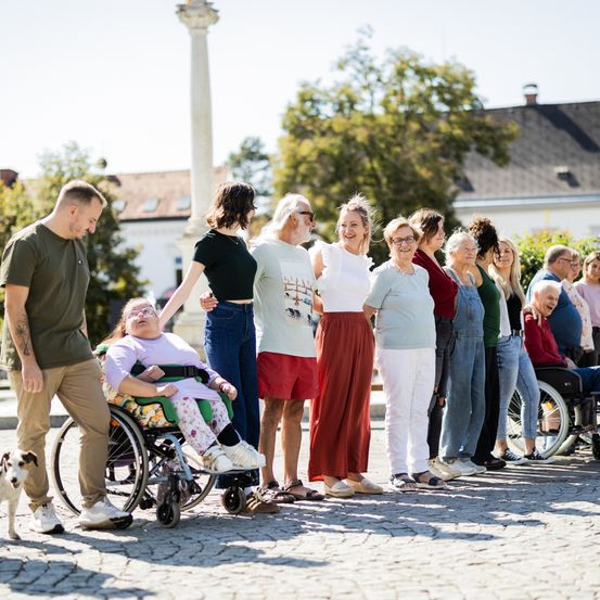 Eine Gruppe von Menschen, darunter einige in Rollstühlen, steht an einem sonnigen Tag vor einem Gebäude. Bäume und eine Statue sind im Hintergrund zu sehen.