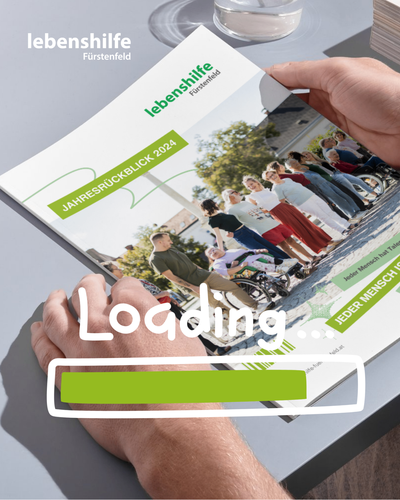 Eine Person hält ein Magazin mit dem Titel 'Lebenshilfe Fürstenfeld Jahresrückblick 2024'. Es zeigt ein Ladesymbol in der unteren rechten Ecke.
