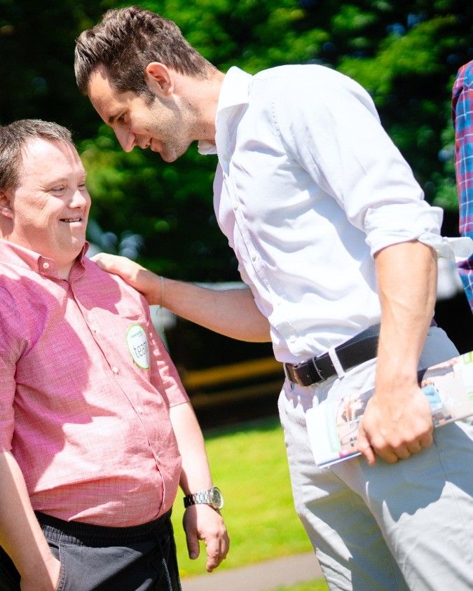 Ein Mann in einem rosa Hemd mit einem grünen Team-Abzeichen lächelt, während ein anderer Mann in einem weißen Hemd seine Schulter hält.