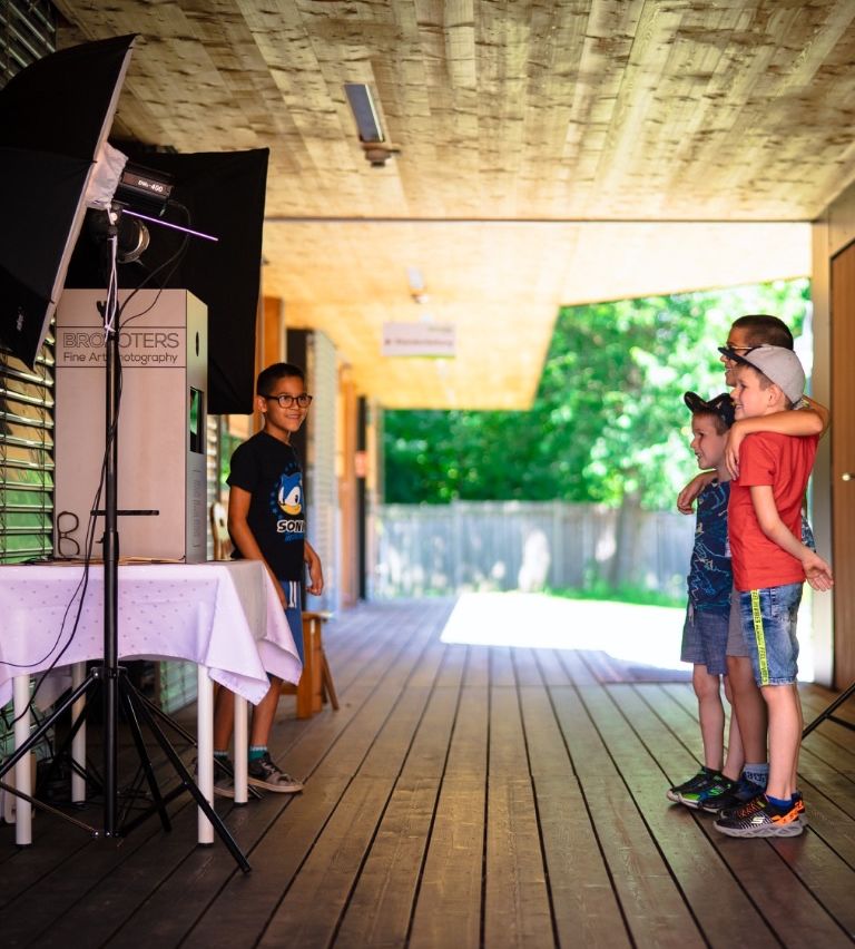 Drei Jungen stehen auf einer hölzernen Veranda mit schwarzem Hintergrund, einer Kamera auf einem Stativ und einem Tisch mit weißer Tischdecke. Ein Junge trägt eine Brille und ein schwarzes T-Shirt mit einem Sonic the Hedgehog-Design.