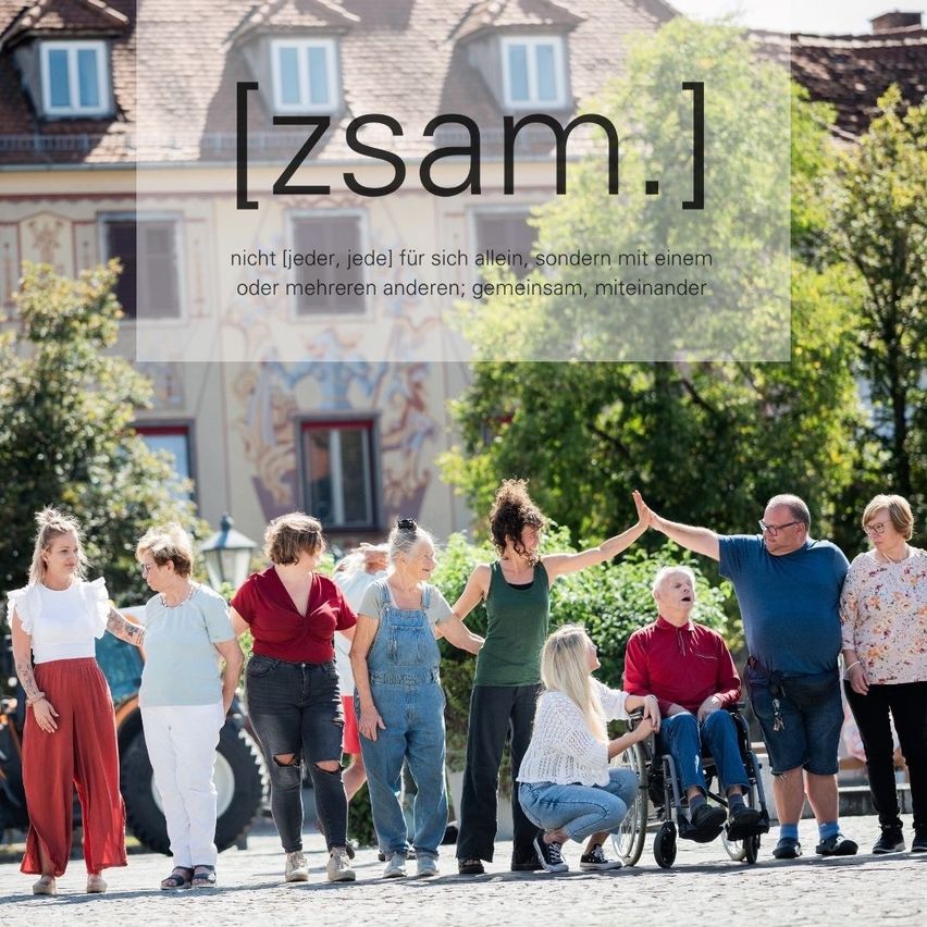 Eine Gruppe von Menschen steht vor einem Gebäude mit einem großen Text, der [zsam.] lautet. Sie alle lächeln und scheinen miteinander zu interagieren.