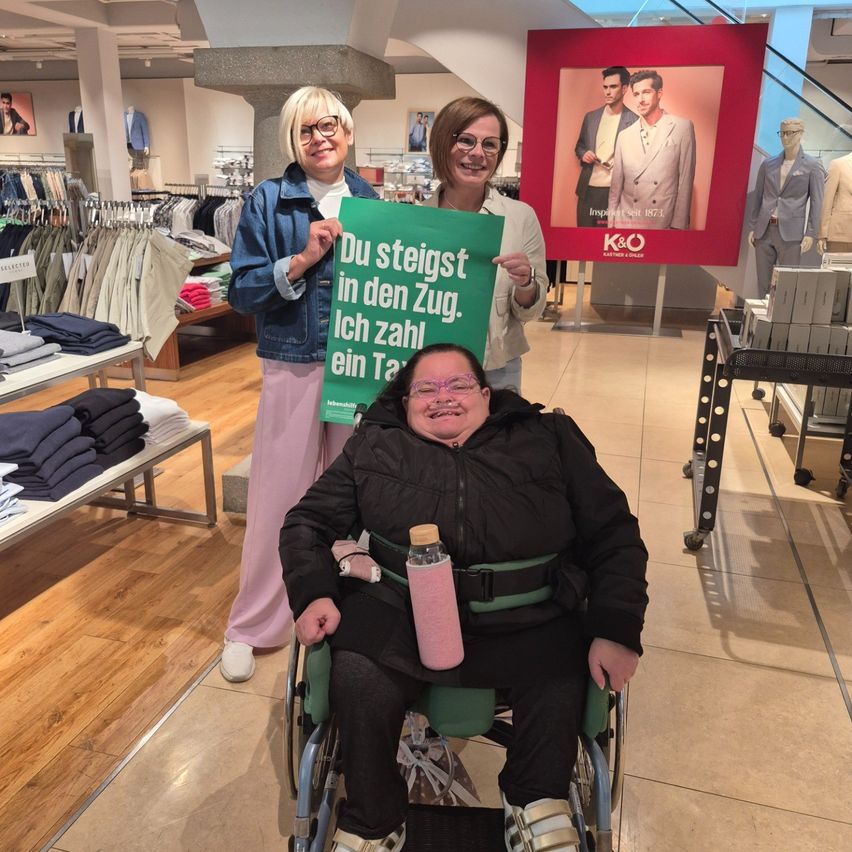 Zwei Frauen stehen hinter einer Frau im Rollstuhl und halten ein grünes Schild hoch. Die Frau im Rollstuhl trägt eine pinke Wasserflasche.