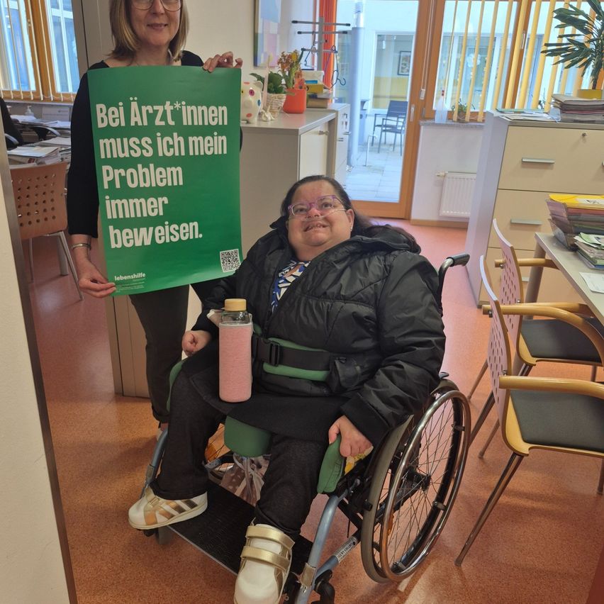 Eine Frau hält ein grünes Plakat mit der Aufschrift: 'Bei Ärzten muss ich mein Problem immer beweisen.' Sie steht neben einer Frau im Rollstuhl.