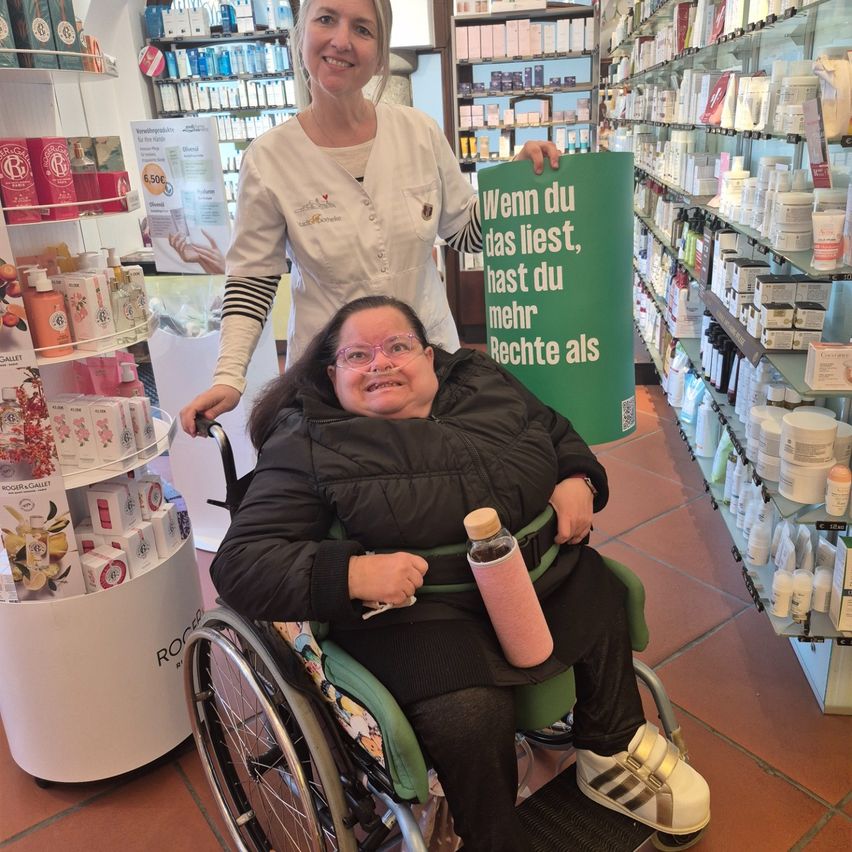 Eine Frau im Rollstuhl mit einem rosafarbenen Becher sitzt vor einer Apotheke, während eine andere Frau hinter ihr ein grünes Plakat hält, auf dem steht: 'Wenn du das liest, hast du mehr Rechte, als du denkst.'