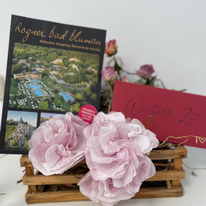 Eine Holzkiste enthält zwei rosa Papierblumen und eine rote Karte mit der Aufschrift 'Wertvolle Zeit'. Dahinter befindet sich eine Broschüre mit dem Titel 'Rogner Bad Blumau'.
