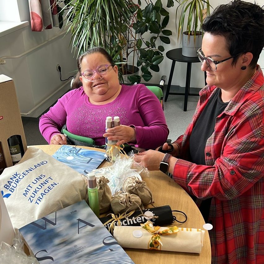 Zwei Frauen, eine im Rollstuhl, lächeln und betrachten Geschenkartikel auf einem Tisch. Sie haben Flaschen und verpackte Geschenke.