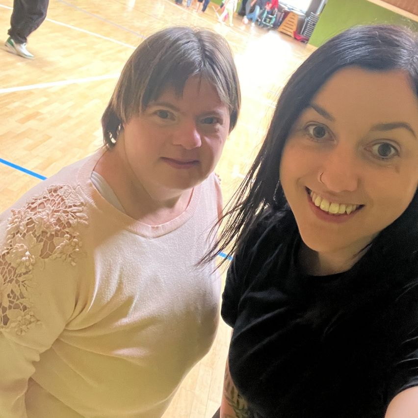 Zwei Frauen lächeln für ein Selfie in einer Turnhalle. Die Frau auf der linken Seite trägt ein rosa Oberteil mit Spitze. Die Frau auf der rechten Seite hat einen Nasenring und ein schwarzes Oberteil.