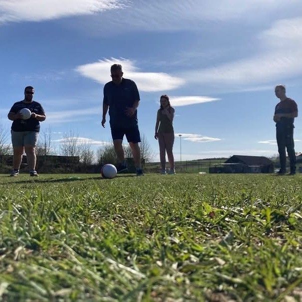 Vier Personen befinden sich auf einem Rasenfeld unter einem blauen Himmel. Ein Mann hält einen Ball und ein anderer ist dabei, ihn zu treten. Eine Frau schaut zu, und ein weiterer Mann steht in der Nähe.