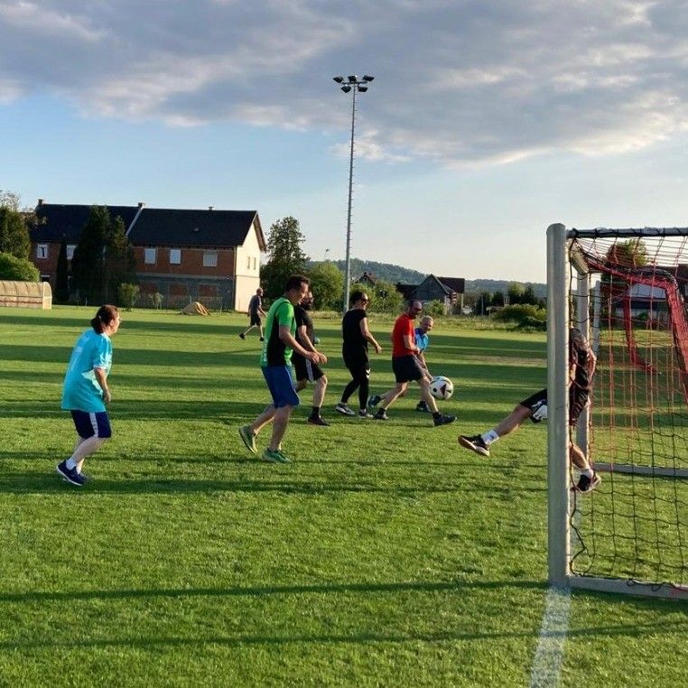 Eine Gruppe von Menschen spielt Fußball auf einem Feld mit bewölktem Himmel und Häusern im Hintergrund.