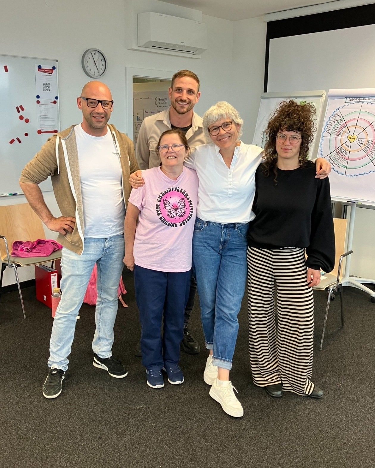 Fünf Personen stehen in einem Klassenzimmer vor einer Tafel. Drei Frauen und zwei Männer lächeln.