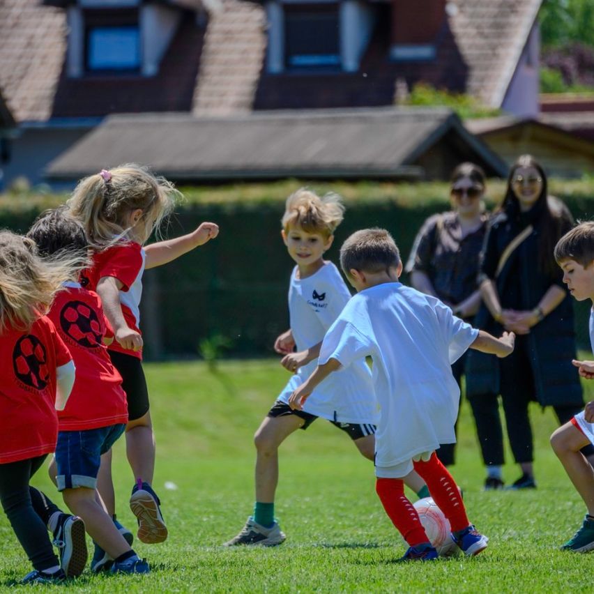 Mehrere Kinder in Fußballtrikots spielen auf einem Rasenplatz, während zwei Frauen hinter einem Zaun zusehen. Ein Kind tritt einen Ball, und ein anderes Kind läuft darauf zu.
