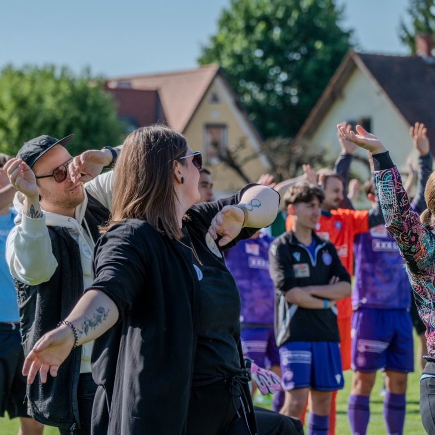 Eine Gruppe von Menschen auf einem Rasenfeld. Eine Frau mit Tätowierungen tanzt, während ein Mann neben ihr ihre Taille hält. Hinter ihnen stehen mehrere Personen und heben ihre Hände. Im Hintergrund sind einige Häuser zu sehen.