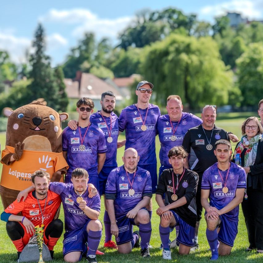 Eine Gruppe von Fußballspielern in lila Trikots mit Medaillen posiert für ein Foto mit einem Maskottchen und Zuschauern in einem Park.
