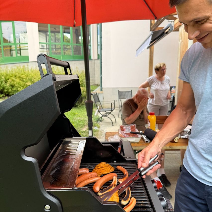 Ein Mann grillt Würstchen und Gemüse auf einem Grill, unter einem roten Schirm. Im Hintergrund sitzt ein älteres Paar am Tisch, wobei eine Person ihr Gesicht bedeckt.