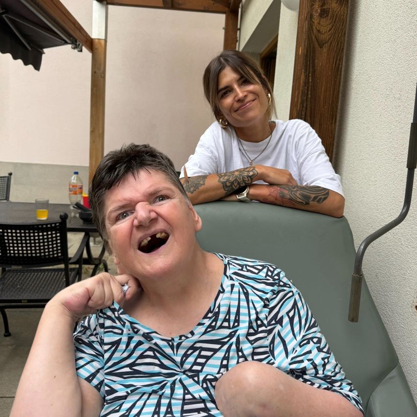Zwei Frauen sitzen auf einer Terrasse, eine lächelt breit mit einer Hand am Kinn, die andere hat Tattoos an den Armen, beide mit einem Tisch und Getränken dahinter.