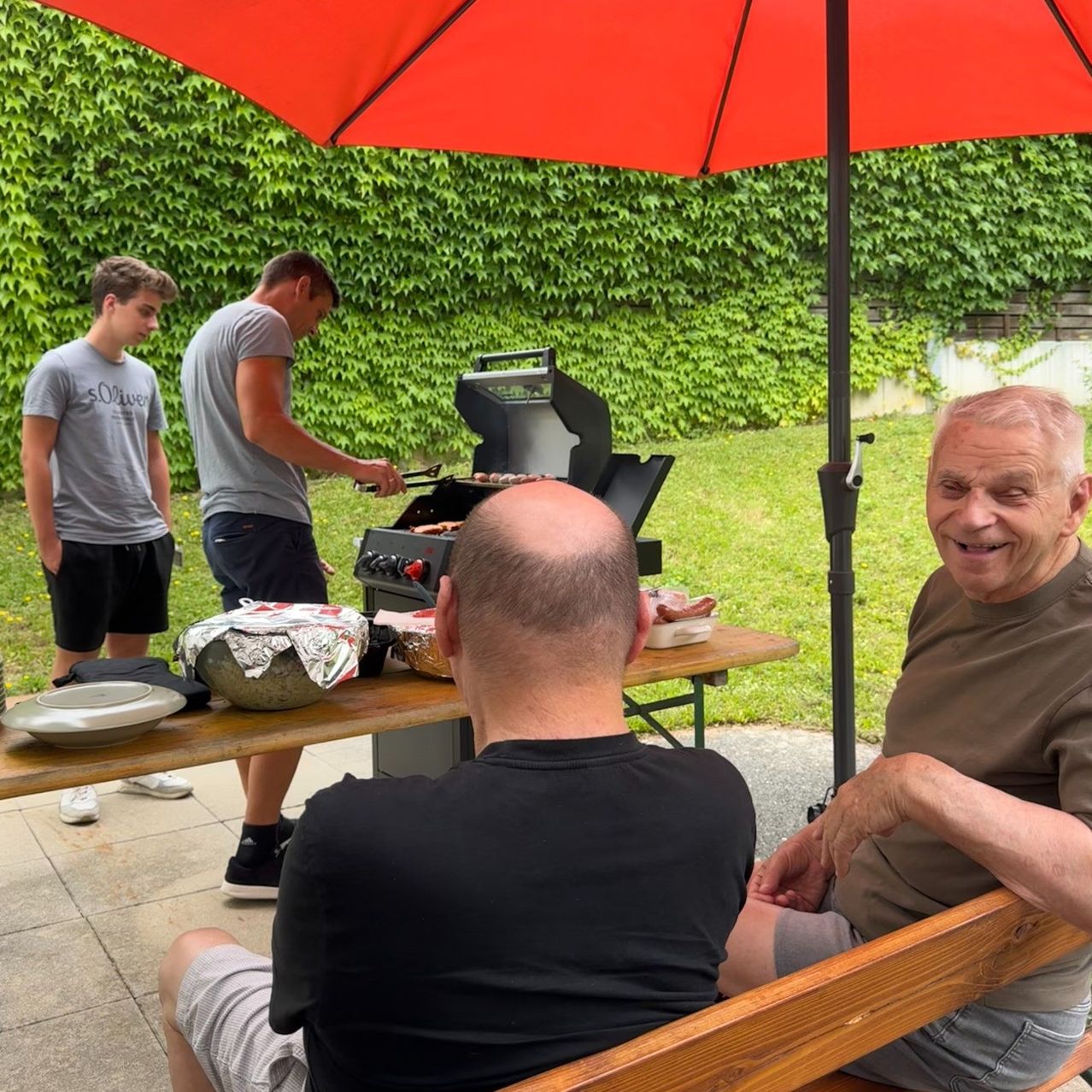Zwei Männer sitzen auf einer Bank unter einem Regenschirm und beobachten einen Mann, der Essen grillt. Der Grill steht auf einem Tisch mit Essen und Tellern. Ein Junge steht hinter dem Grill. Das Gelände ist mit grüner Efeu bedeckt.