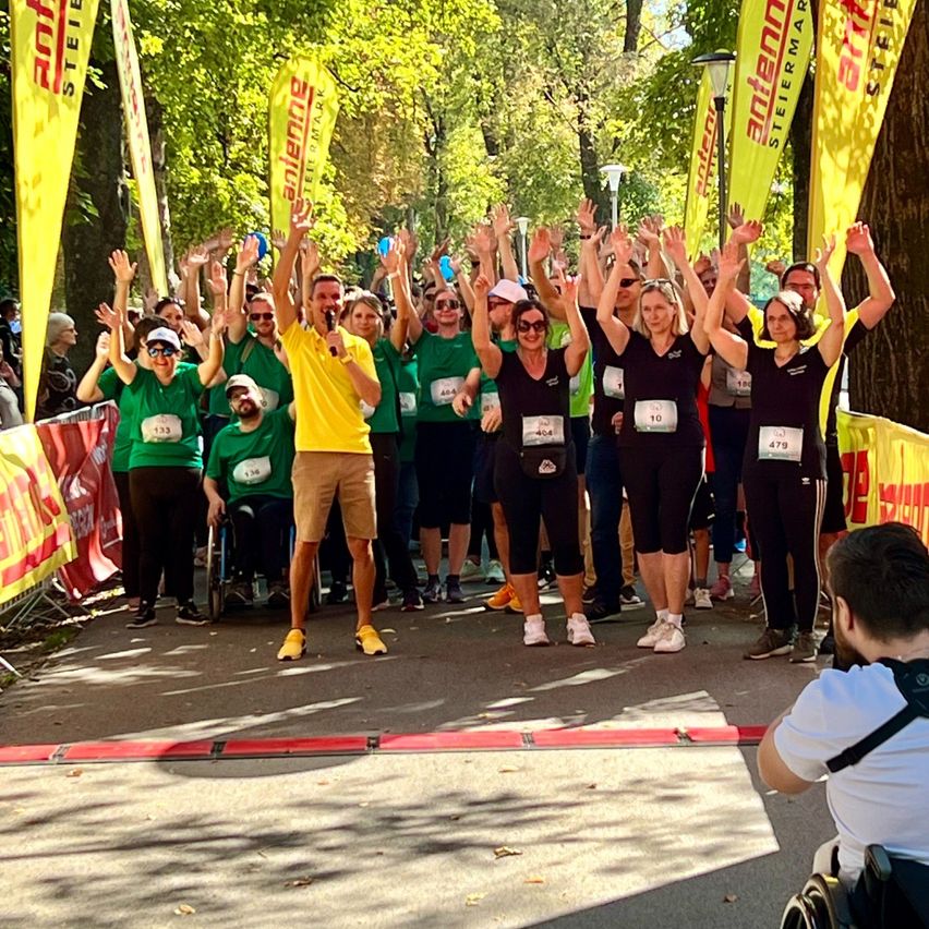 Eine Gruppe von Läufern in grünen Shirts versammelt sich, hebt die Hände, ein Mann in Gelb steht vor ihnen. Sie sind von Bäumen und Bannern umgeben.