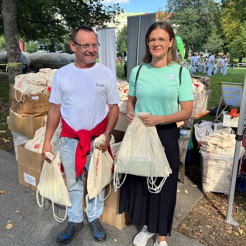 Ein erwachsener Mann und eine Frau stehen nebeneinander und halten Taschen. Sie befinden sich in einem Außenbereich mit Bäumen und Menschen im Hintergrund. Der Mann trägt eine Brille und einen roten Umhang. Die Frau trägt ein grünes T-Shirt und eine schwarze Rock. Hinter ihnen befinden sich Kisten und Taschen.
