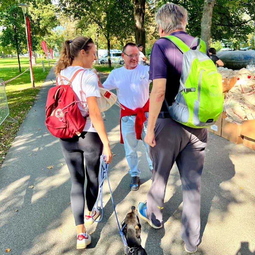 Drei Personen gehen auf einem Pfad in einem Park. Eine Frau führt einen kleinen Hund. Ein Mann in der Mitte trägt ein rotes Band und lächelt. Ein Mann auf der rechten Seite trägt einen Rucksack. Bäume und Fahnen säumen den Weg.