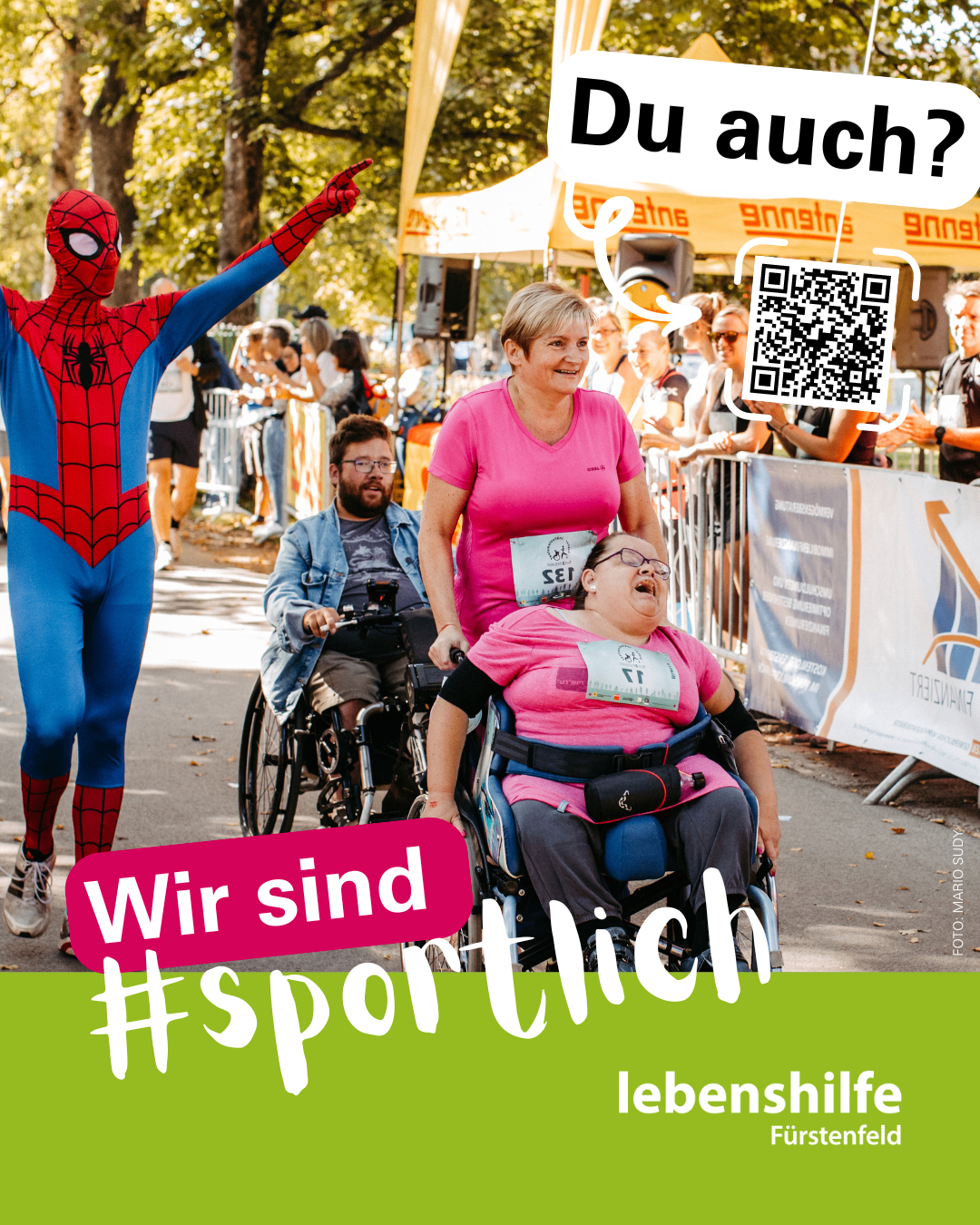 Eine Frau im Rollstuhl wird von einer anderen Frau geschoben. Ein Mann in einer Spiderman-Kostümierung steht neben ihnen. Sie befinden sich in einem Rennen. Die Frau im Rollstuhl hat eine Nummer auf ihrem Shirt. In der Ecke befindet sich ein QR-Code.