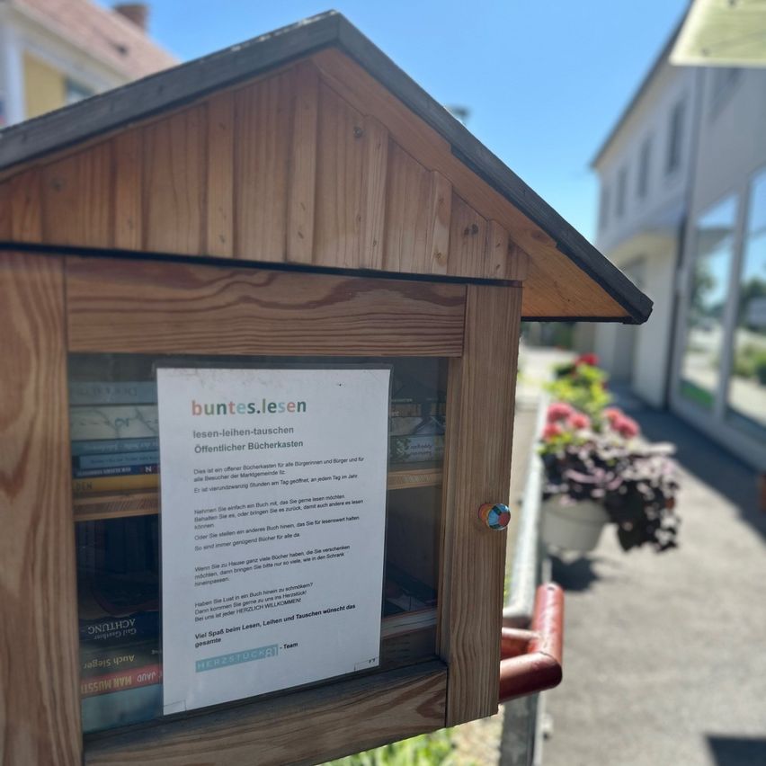 Eine hölzerne Büchertauschbox steht auf dem Bürgersteig mit einem Schild, das 'bunte.lesen' sagt. Sie enthält Bücher und steht neben einem Gebäude mit Blumen.