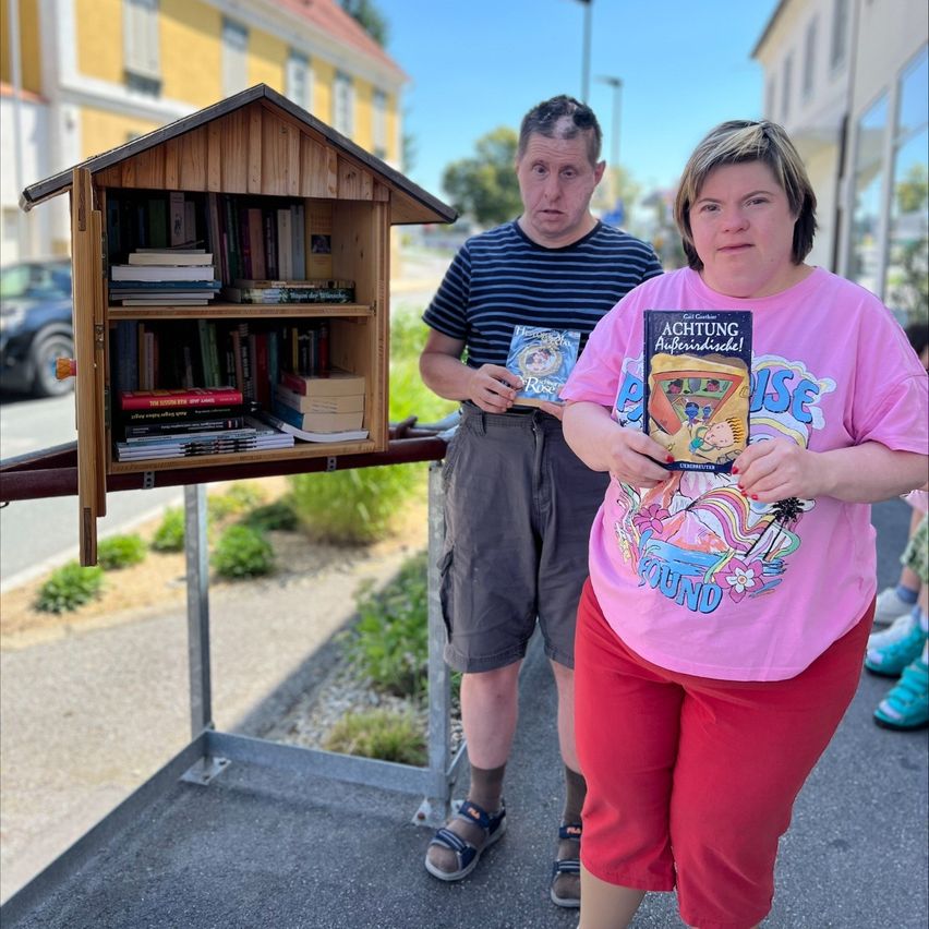Zwei Personen stehen neben einer kleinen hölzernen Bibliotheksbox, die mit Büchern gefüllt ist, an einem sonnigen Tag. Der Mann hält zwei Bücher, und die Frau hält eines. Sie tragen beide Freizeitkleidung. Hinter ihnen steht ein Gebäude mit einer gelben Wand.