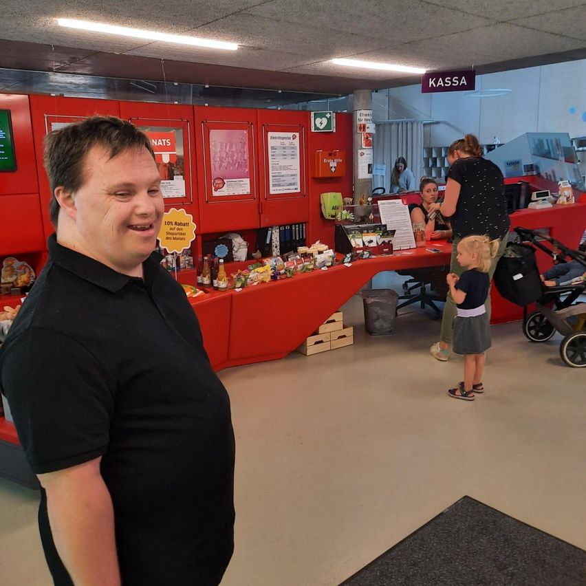 Ein Mann steht lächelnd vor der Kamera in einem Geschäft, mit einer Theke und einem Schild, das 'Kassa' in der Hintergrund steht. Hinter ihm steht ein kleines Kind, und eine Frau bedient die Theke.