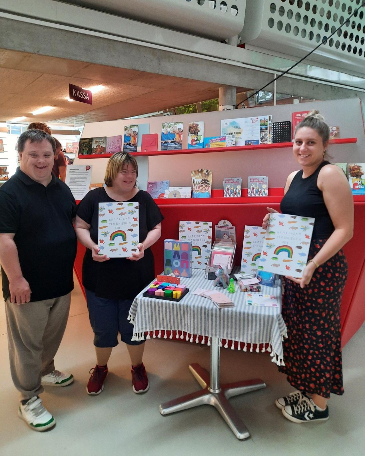 Drei Menschen lächeln in einer Bibliothek mit einem Tisch voller Bücher und Gegenständen. Eine Frau auf der rechten Seite hält ein buntes Buch. Ein Mann auf der linken Seite trägt ein schwarzes T-Shirt.