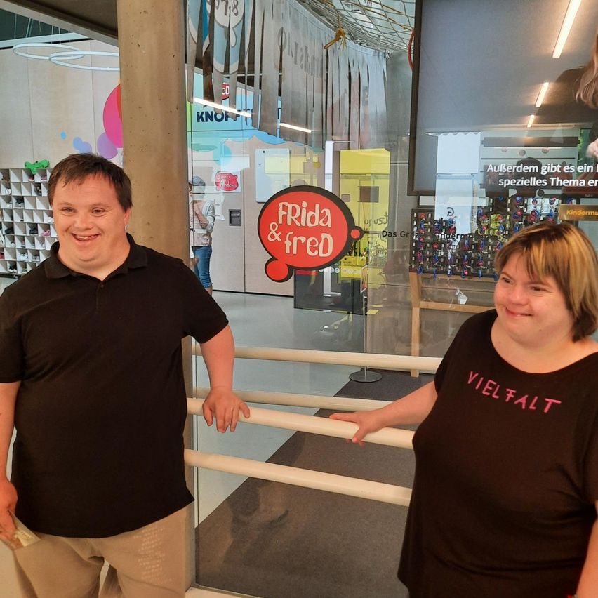 Zwei Menschen in schwarzen T-Shirts stehen vor einer Glastür mit den Worten 'FRIDA & FRED'. Der Mann lächelt und hält Geld, während die Frau lächelt und posiert. Der Hintergrund zeigt einen verschwommenen Innenraum mit Regalen und einem großen Bildschirm.