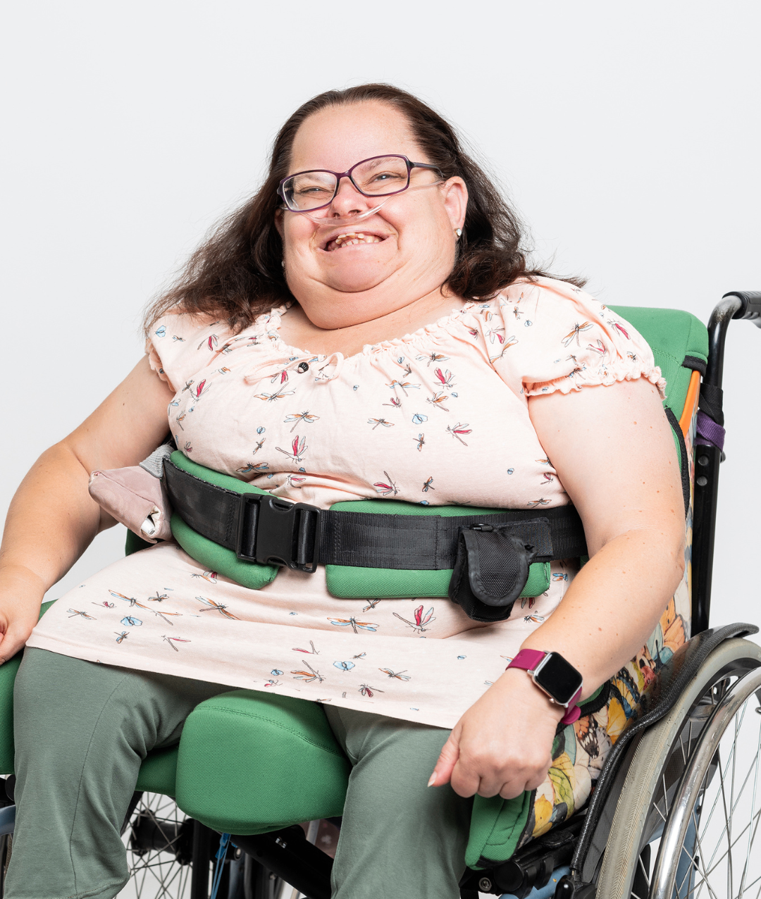 Eine Frau im Rollstuhl lächelt, trägt eine Brille und ein rosa Kleid mit Drachenfliegenmuster. Sie hat einen schwarzen Sicherheitsgurt um die Taille und eine Smartwatch am Handgelenk.