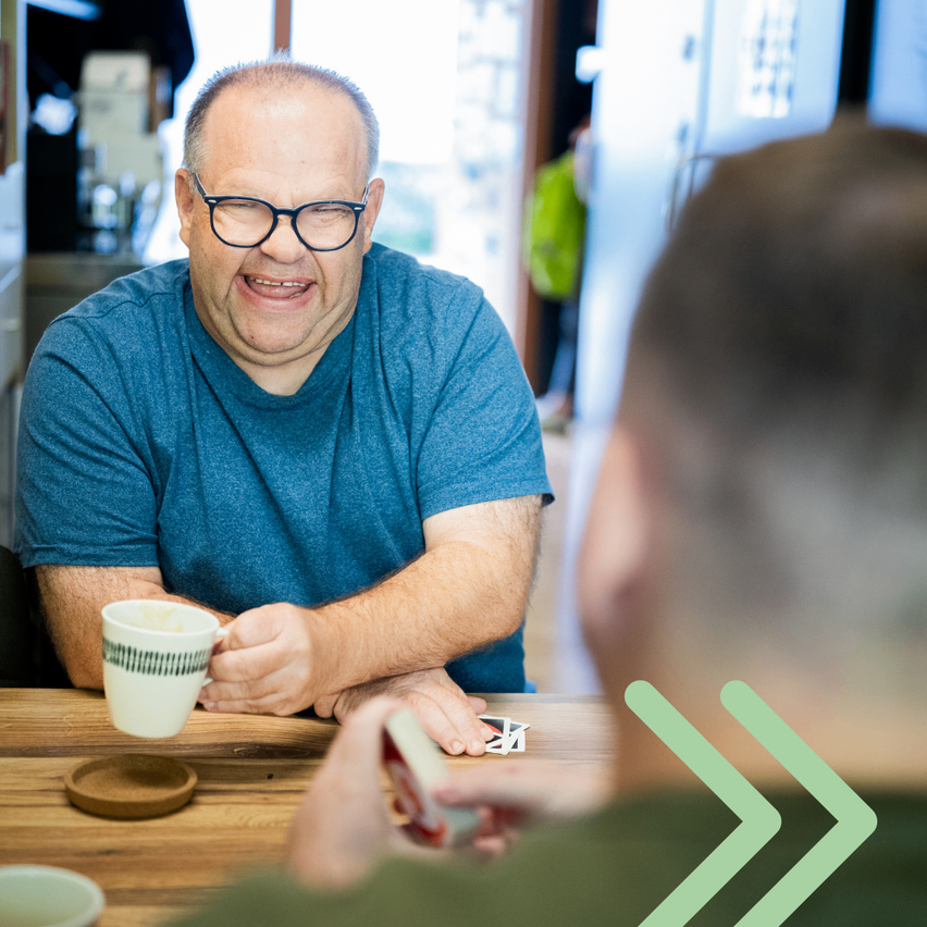 Ein Mann mit Brille und blauem T-Shirt lächelt, während er eine Kaffeetasse auf einem Holztisch hält. Ein anderer Mann in einem grünen Hemd schaut ihn an.