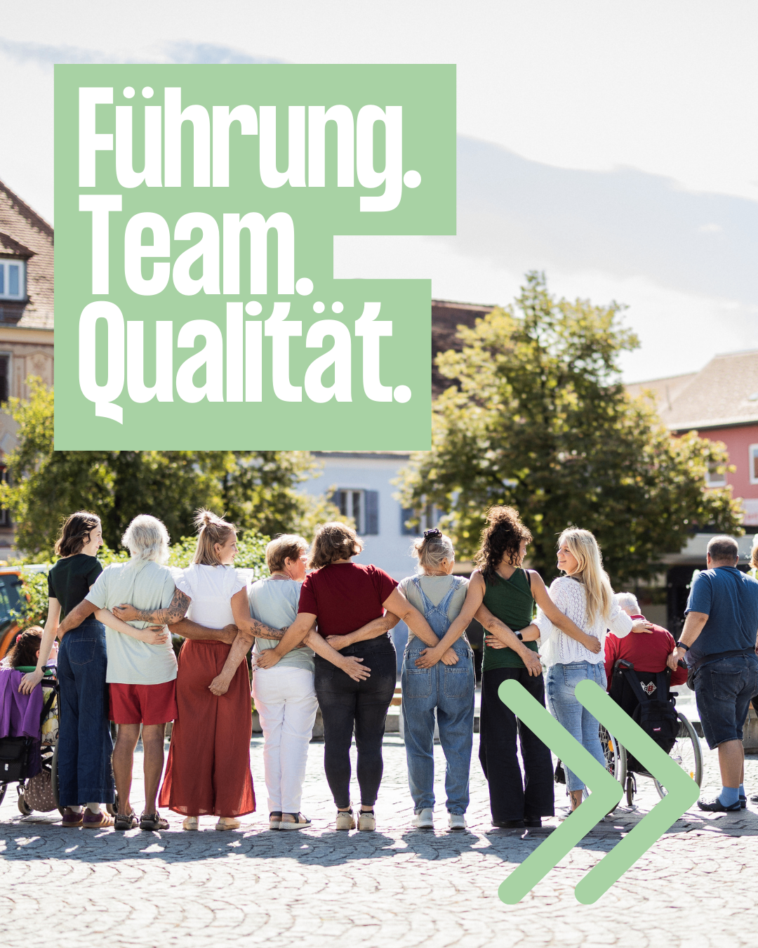 Eine Gruppe von Menschen, darunter ältere Personen und Menschen im Rollstuhl, stehen eng beieinander und haben ihre Arme umeinander geschlungen. Im Hintergrund befindet sich ein grünes Schild mit den Worten 'Führung. Team. Qualität.' und ein bewölkter Himmel.