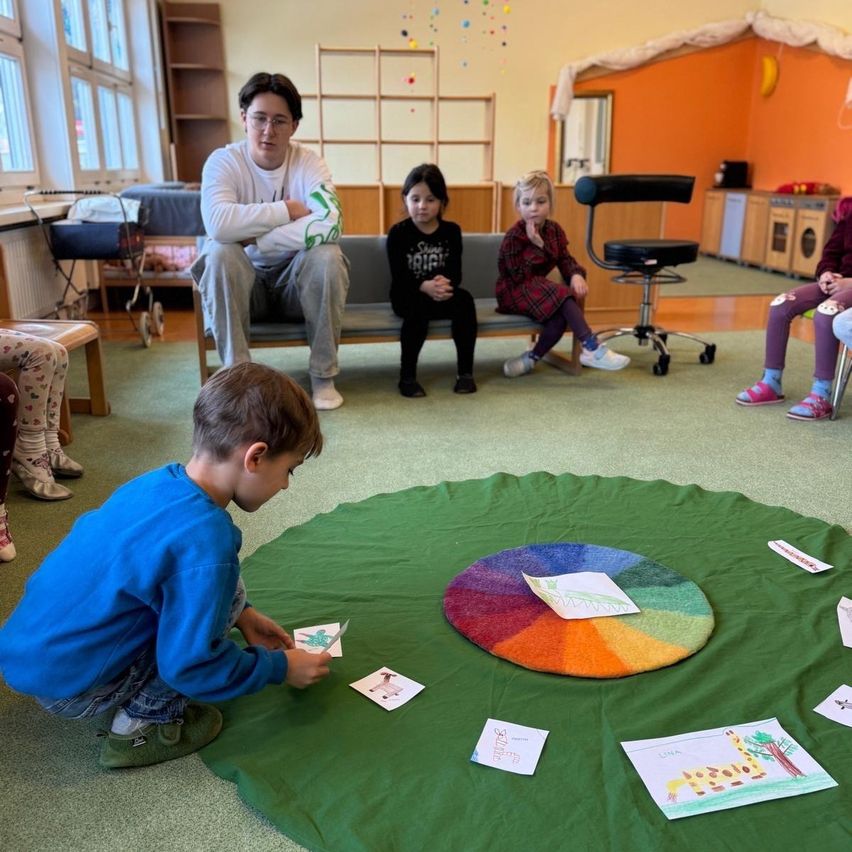 A boy places cards on a color wheel in a playroom. Several children sit around the wheel, observing. An adult sits on a bench nearby.