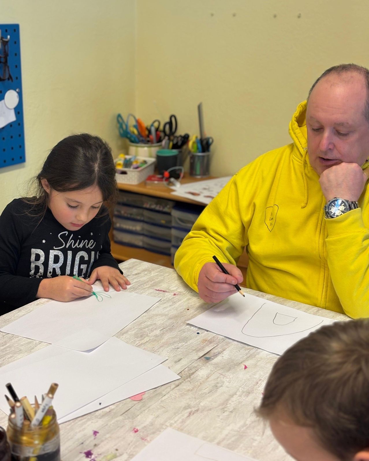 A man and a girl are drawing on a desk. The girl is wearing a black sweatshirt and the man is in a yellow hoodie. They are both focused on their drawings. There are pencils, markers, and papers on the desk. In the background, there is a desk with various items.