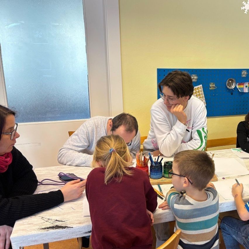 A group of people, including children, are seated around a table in a classroom, engaged in an activity. They have papers, pens, and various art supplies on the table.
