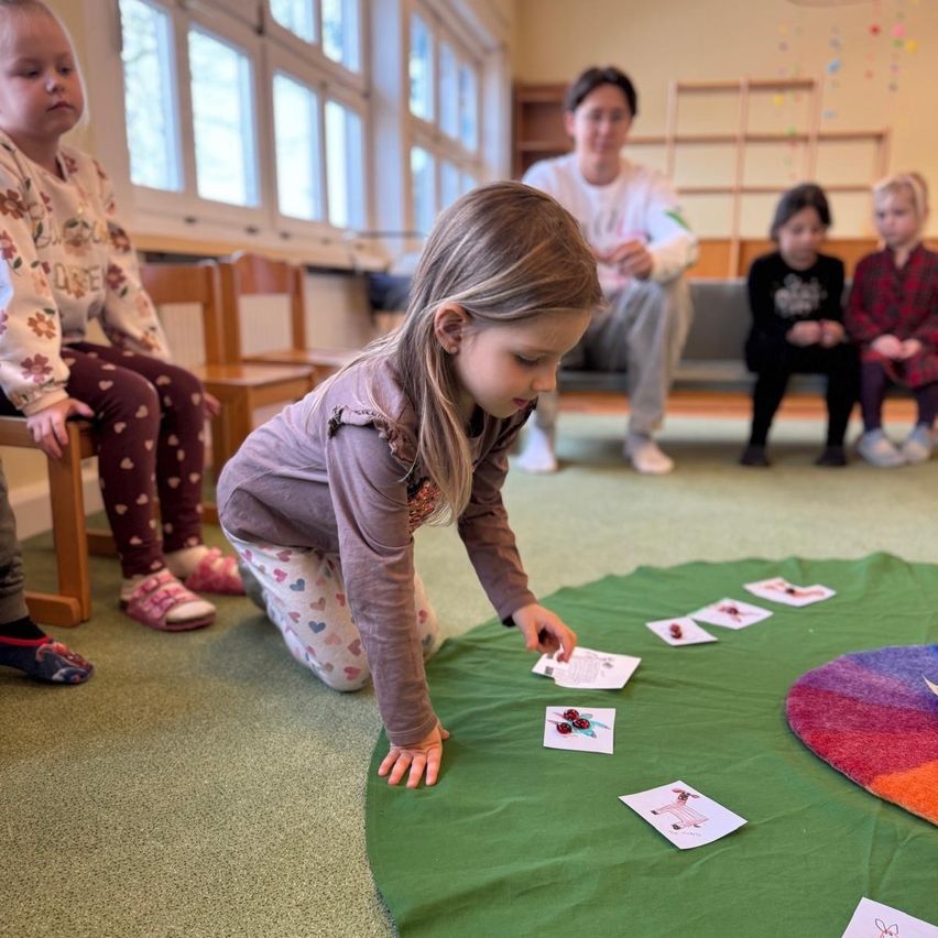 A young girl plays with cards on a colorful rug while a few children and an adult watch in a room with large windows.