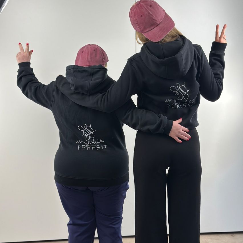 Two women wearing black hoodies with white prints stand back-to-back, one with a peace sign, against a white background.
