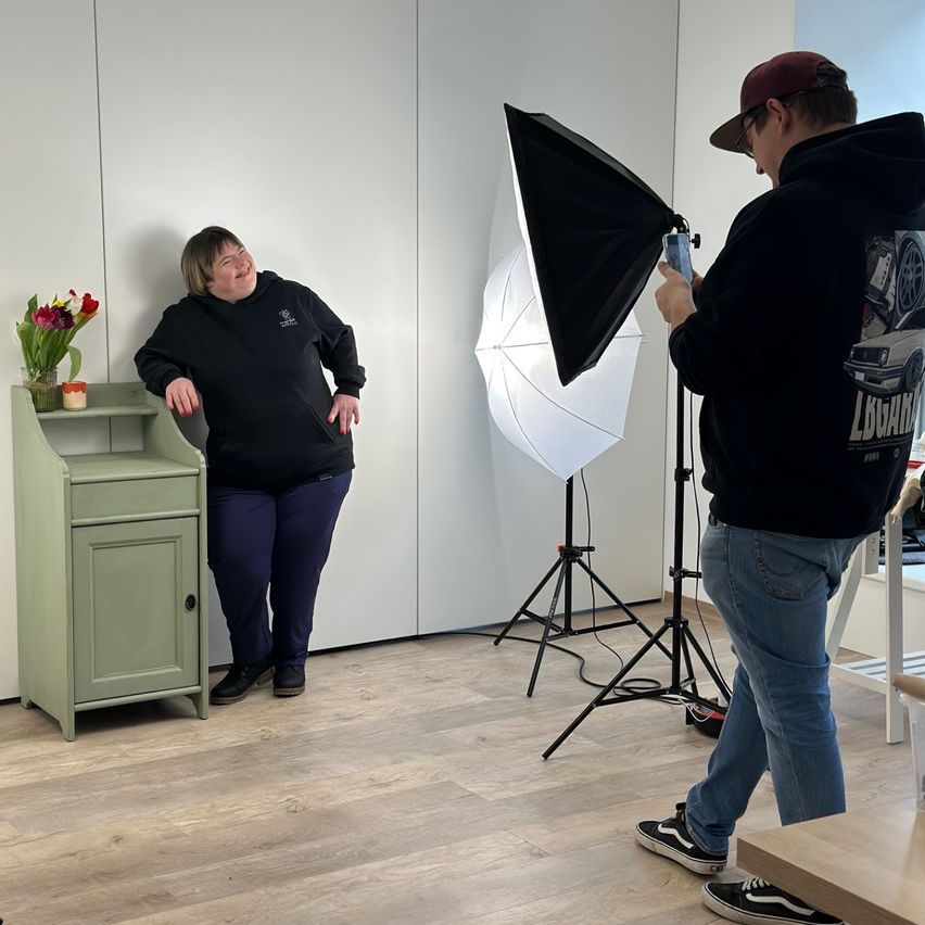A woman in black clothing smiles near a green cabinet while a man takes a photo with a camera and a flash.