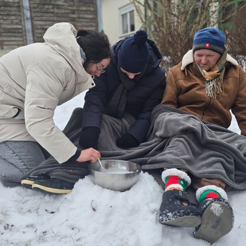 Three people sit on the snowy ground, preparing a meal. One person stirs a bowl while the others watch. They are warmly dressed, with scarves and winter boots.