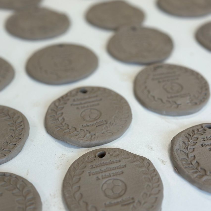 Eine Sammlung von runden Tonschildchen ist auf einer weißen Oberfläche ausgestellt. Jedes Schildchen hat ein Lorbeerkranzdesign und eingravierten Text. Der Text variiert und enthält Daten und Namen. Die Schildchen sind einheitlich geformt und bemessen.