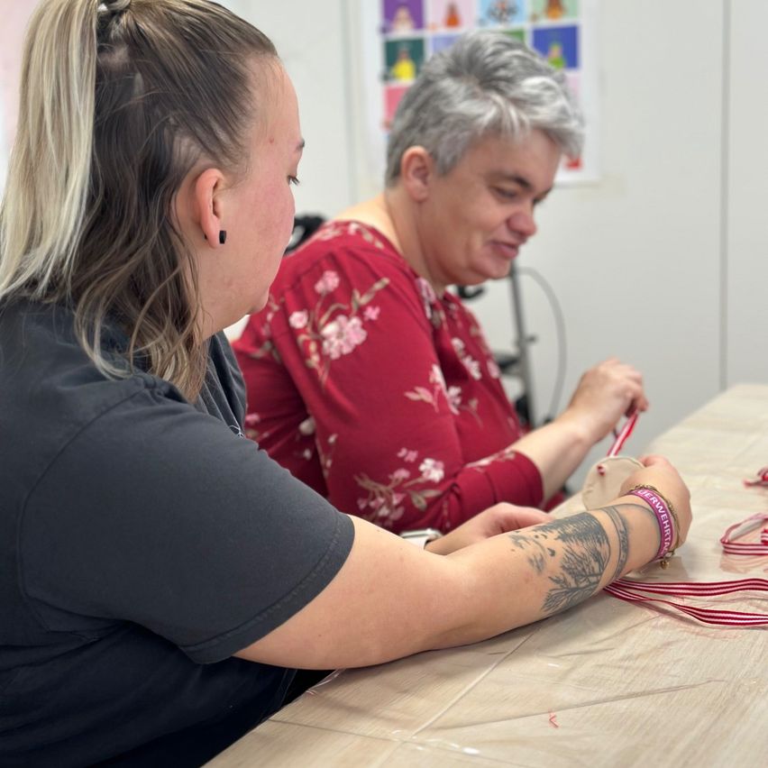Zwei Frauen sitzen an einem Tisch und basteln mit Bändern. Eine Frau lächelt, während die andere zusieht. Im Hintergrund befindet sich ein Blumenmuster und medizinische Geräte.