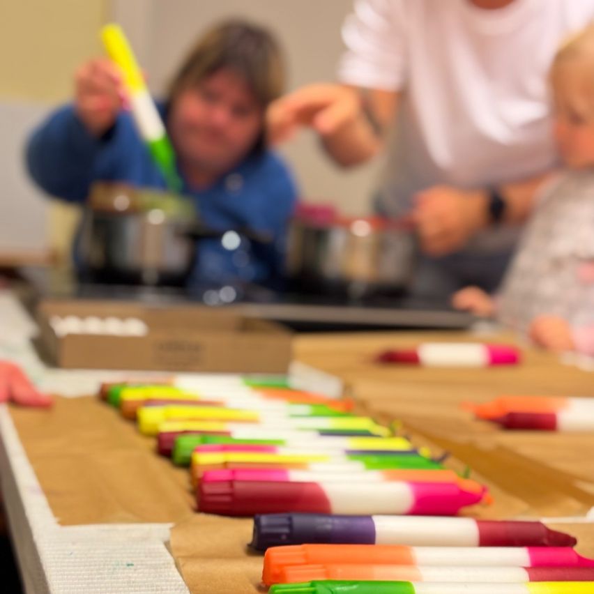 Eine Gruppe von Menschen, darunter eine Frau und ein Kind, versammelt sich um einen Tisch. Sie halten bunte Marker in den Händen und scheinen eine Aktivität durchzuführen.